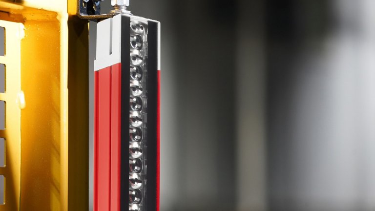 A close-up view of the top 1/3 of a red 450L Guardshield safety light curtain, with grey end cap, side-mounted to a yellow metal machine with a greyed out blurry background. The optical lens side is facing to the right.