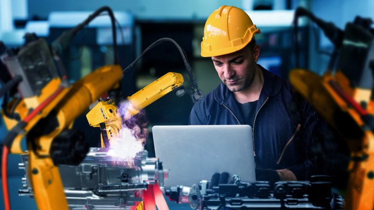 Rockwell Technician in hardhat working on laptop in Modern manufacturing plant floor with industrial robots representing iiOT concept