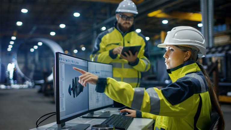 Two workers wearing yellow safety jackets and white hard hats.  The woman is in the foreground pointing to dashboards on a computer screen, the man holding a tablet.  With a manufacturing  space in the background
